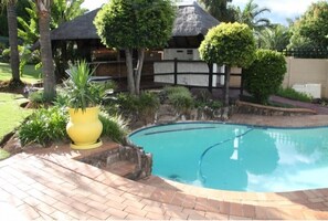 Une piscine entourée d’une terrasse en briques, d’une structure à toit de chaume et d’une végétation luxuriante.