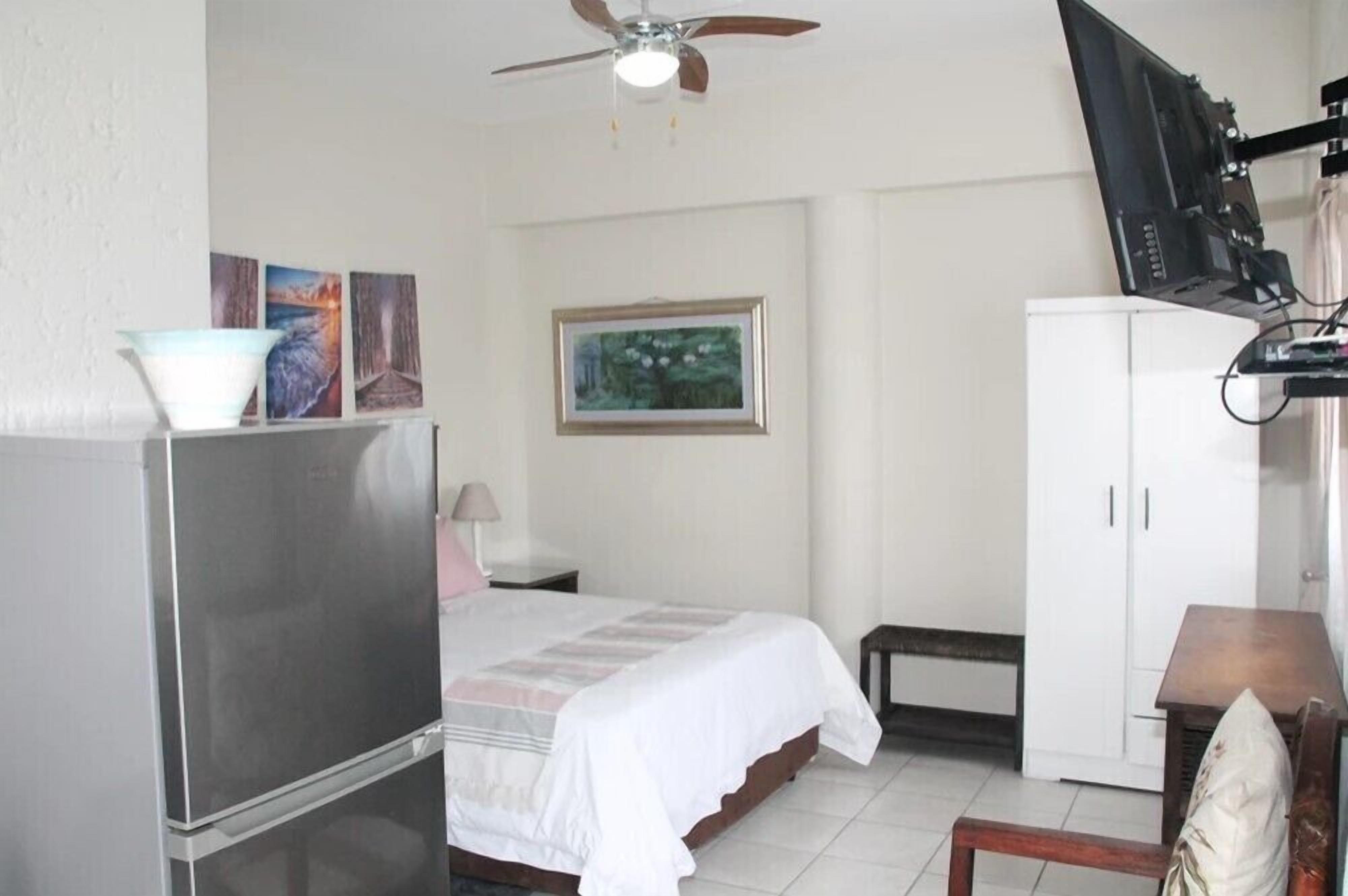 Quarto de hotel compacto com cama, escrivaninha, televisão na parede, ventoinha no teto e armário.