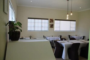 Un espace repas avec des tables préparées pour deux personnes, de grandes fenêtres et des luminaires suspendus.