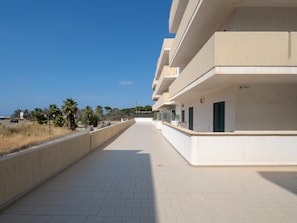 A long, straight pathway leading to a multi-story building with balconies, a clear sky, and palm trees in the background.