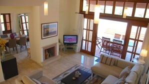 A spacious living room with a sofa, coffee table, and a television. There is a fireplace, dining area, and large windows with curtains. - Villa 32 - Limni (Kouklia)