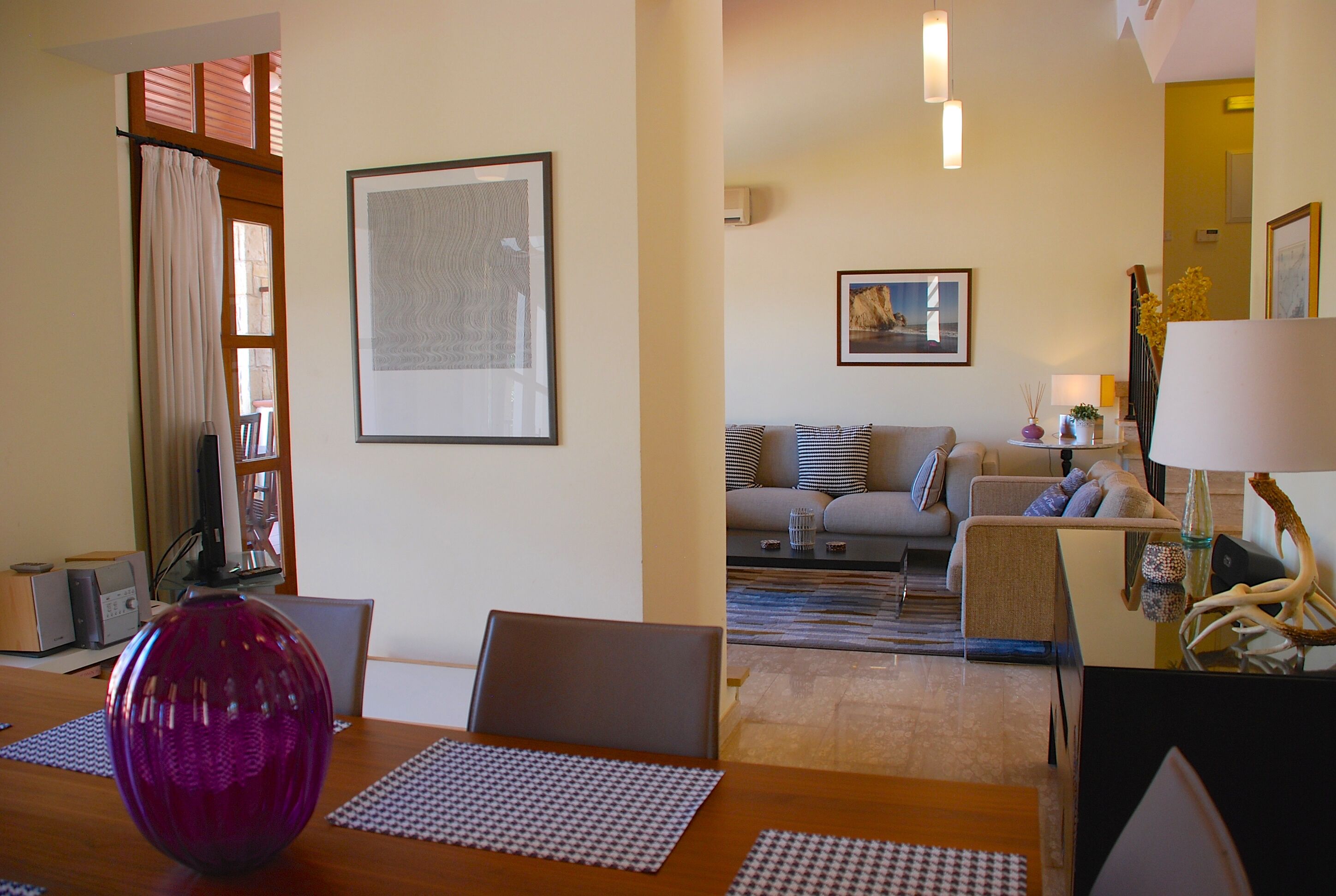 A living room with a sofa set, a dining table with placemats, a large framed abstract artwork, and a decorative vase.