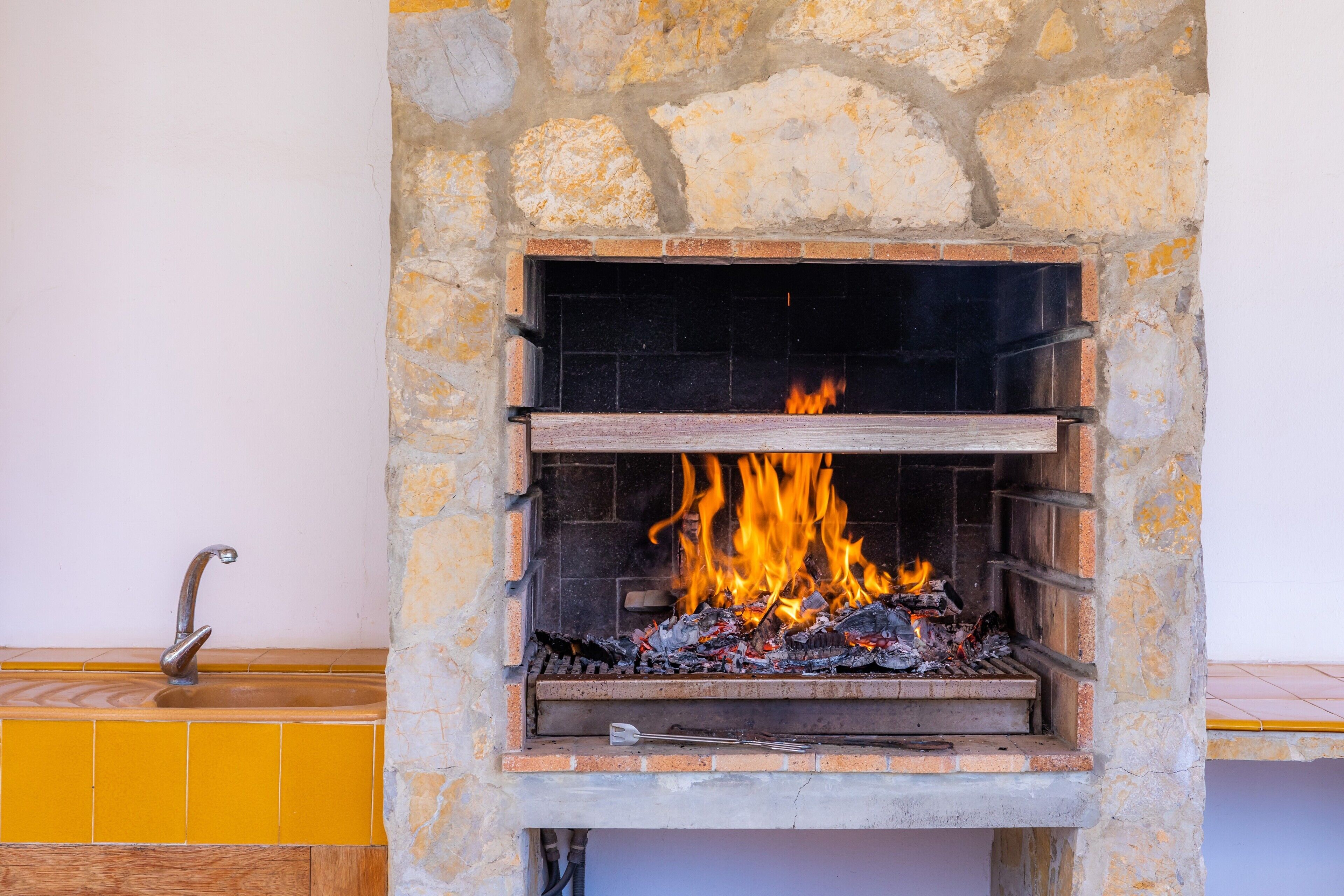 A stone fireplace with a fire burning inside, a wooden shelf below, and a yellow tiled wall to the left.