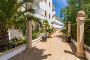 A corridor with potted plants, a palm tree, and white buildings with balconies. - Apartment Close to the Beach with Pool Access, Private Terrace, Air Conditioning and Wi-Fi (Canyamel)