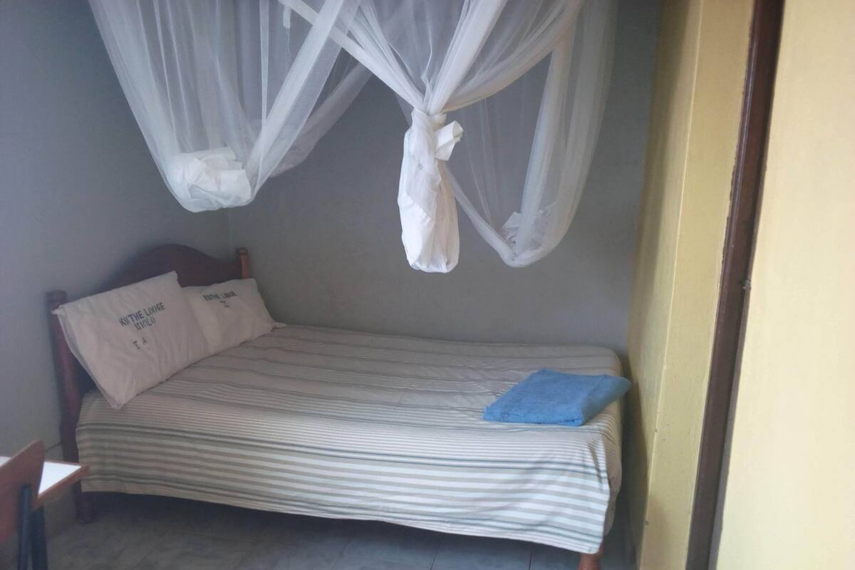 A single bed with a striped bedspread, two pillows, a blue towel, and a mosquito net.