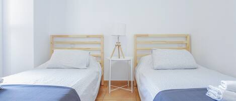 A hotel room with two single beds, a small table with a lamp, and a window.