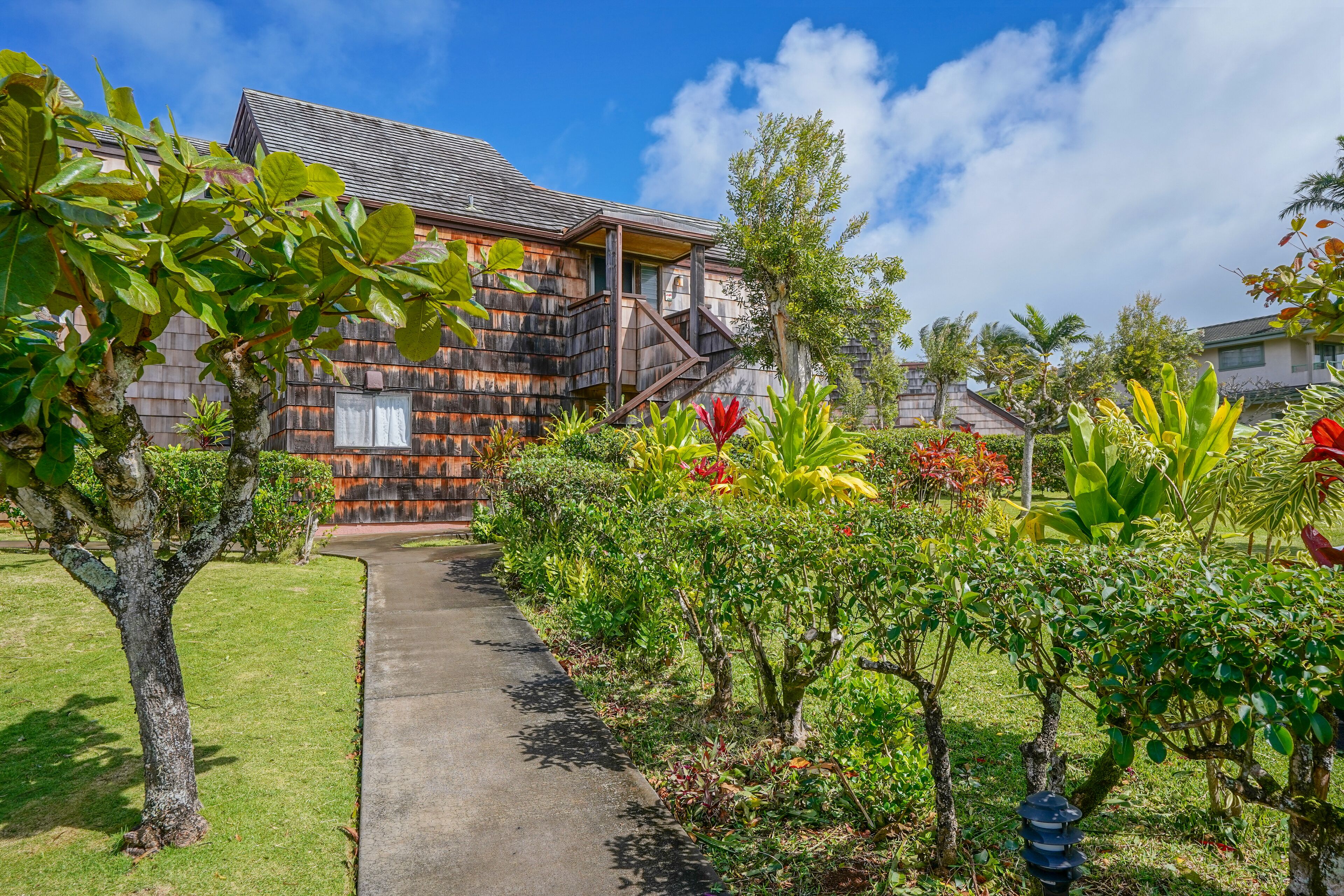 A wooden house with a staircase leading to the upper floor, surrounded by lush greenery and a clear sky.