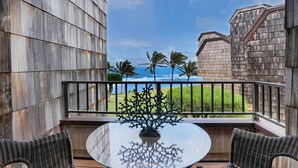A balcony with wicker furniture and a glass table, overlooking a grassy area and palm trees with a view of the ocean. - Sealodge H5: Delightful Oceanview Condo, Close to Beaches & More (Princeville)