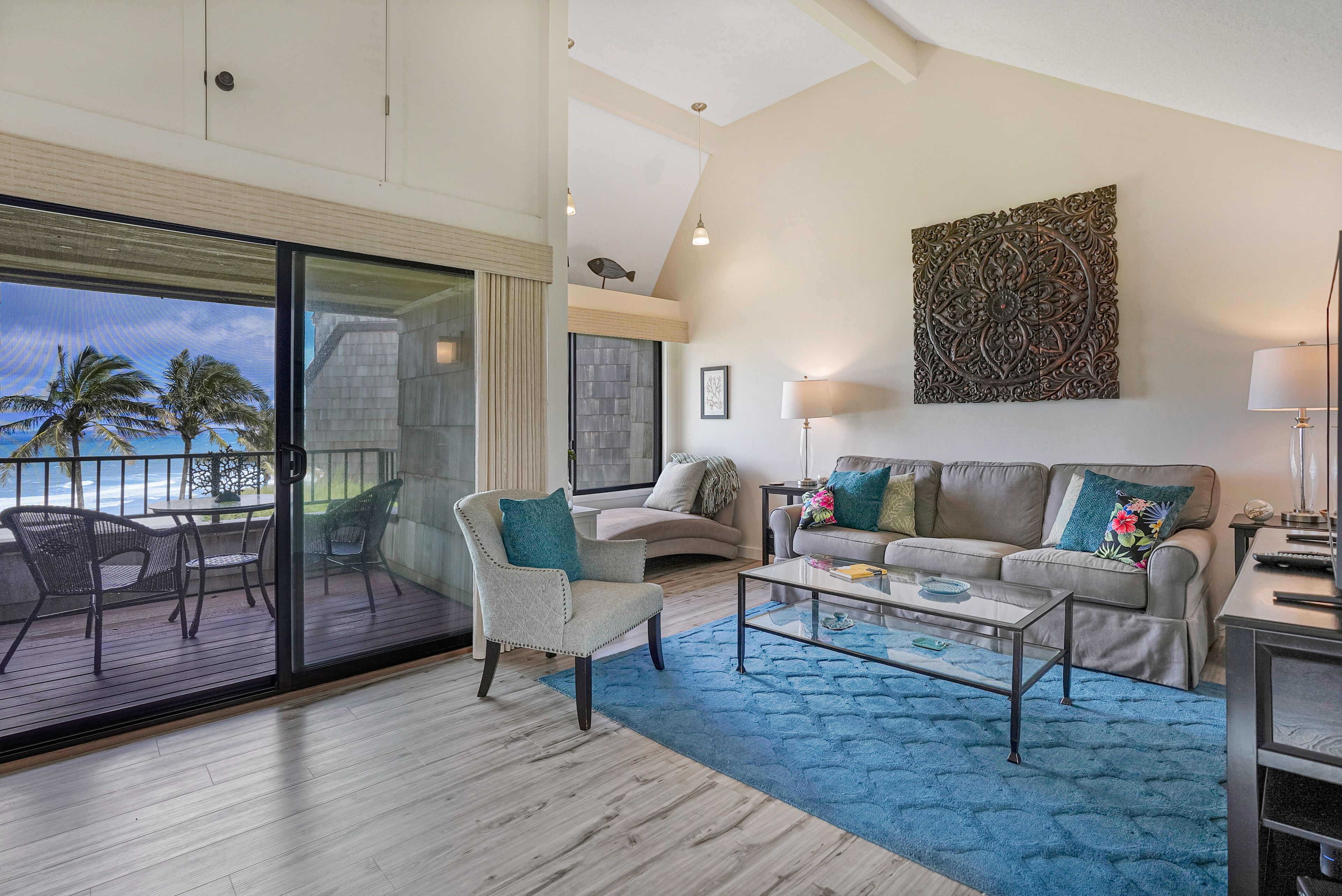 A spacious living room with a large sliding glass door leading to a balcony, a sofa set, a coffee table, and a decorative wall piece.