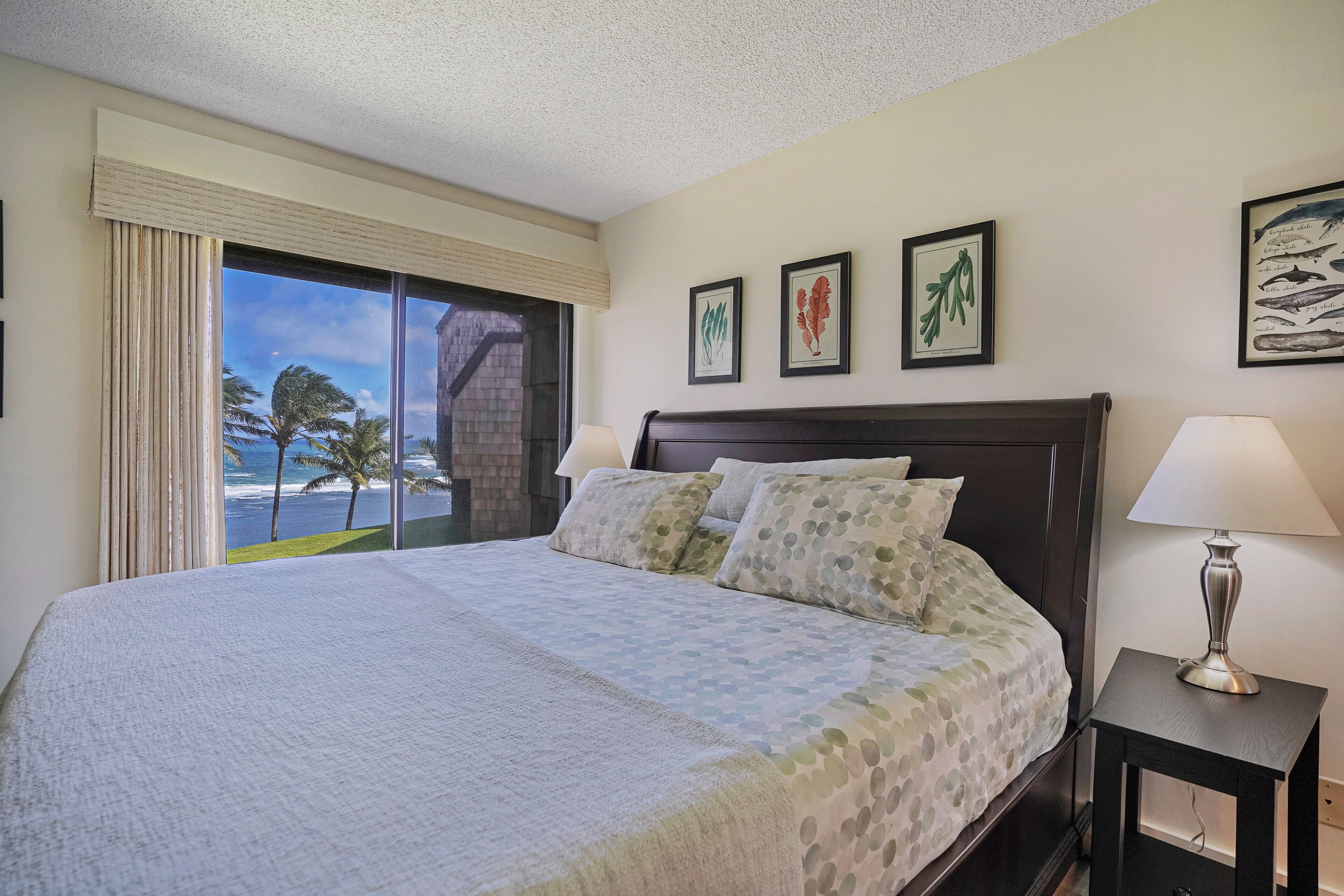 A hotel room with a large bed, two bedside lamps, a window with a view of palm trees and the ocean, and three framed botanical prints on the wall.