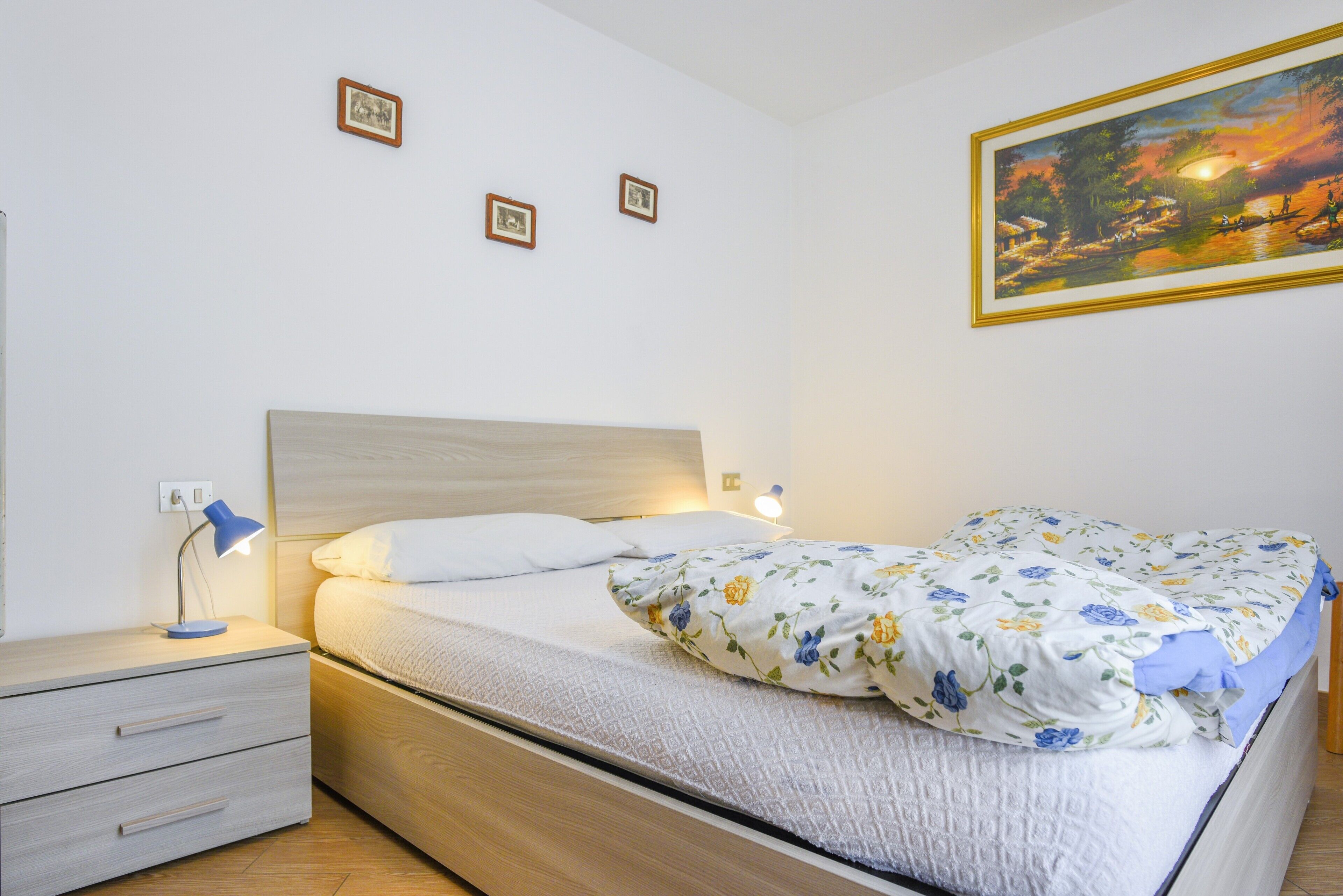 Een slaapkamer met een bed, nachtkastje, lamp en een aan de muur bevestigd schilderij.