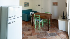 A compact kitchen with a white refrigerator, a wooden dining table with chairs, a teal sofa, and a white cabinet. - Holiday Home 'Il Timo' with Sea View, Wi-Fi and Air Conditioning (Marciana)