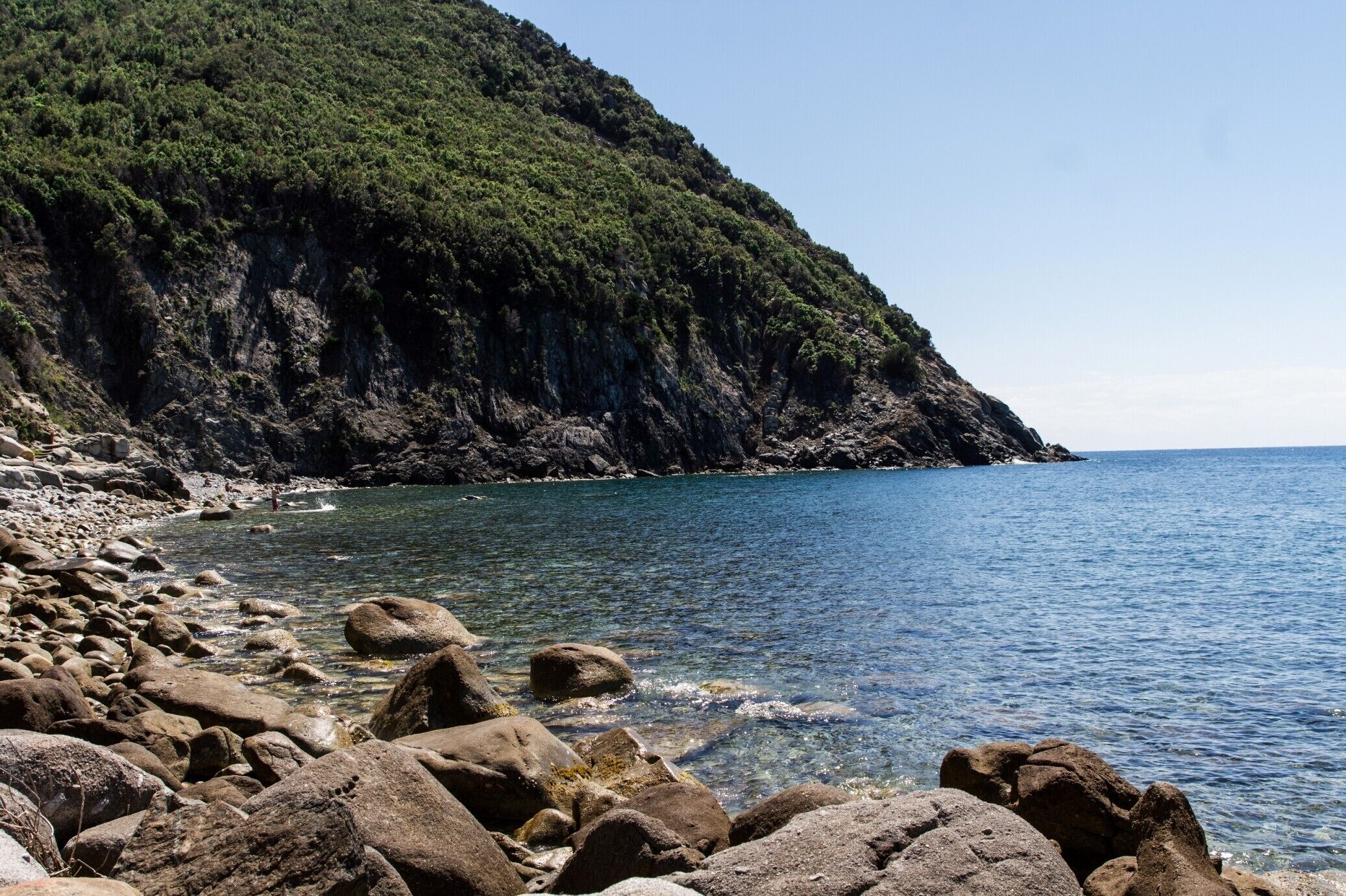 A rocky shoreline with clear water, a steep cliff, and a clear sky.