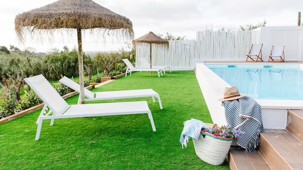 A poolside area with a grassy lawn, white lounge chairs, a thatched umbrella, and a white fence.