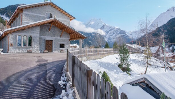Ein Steinhaus mit Holzdach, einem Holzzaun und schneebedeckten Bergen im Hintergrund.