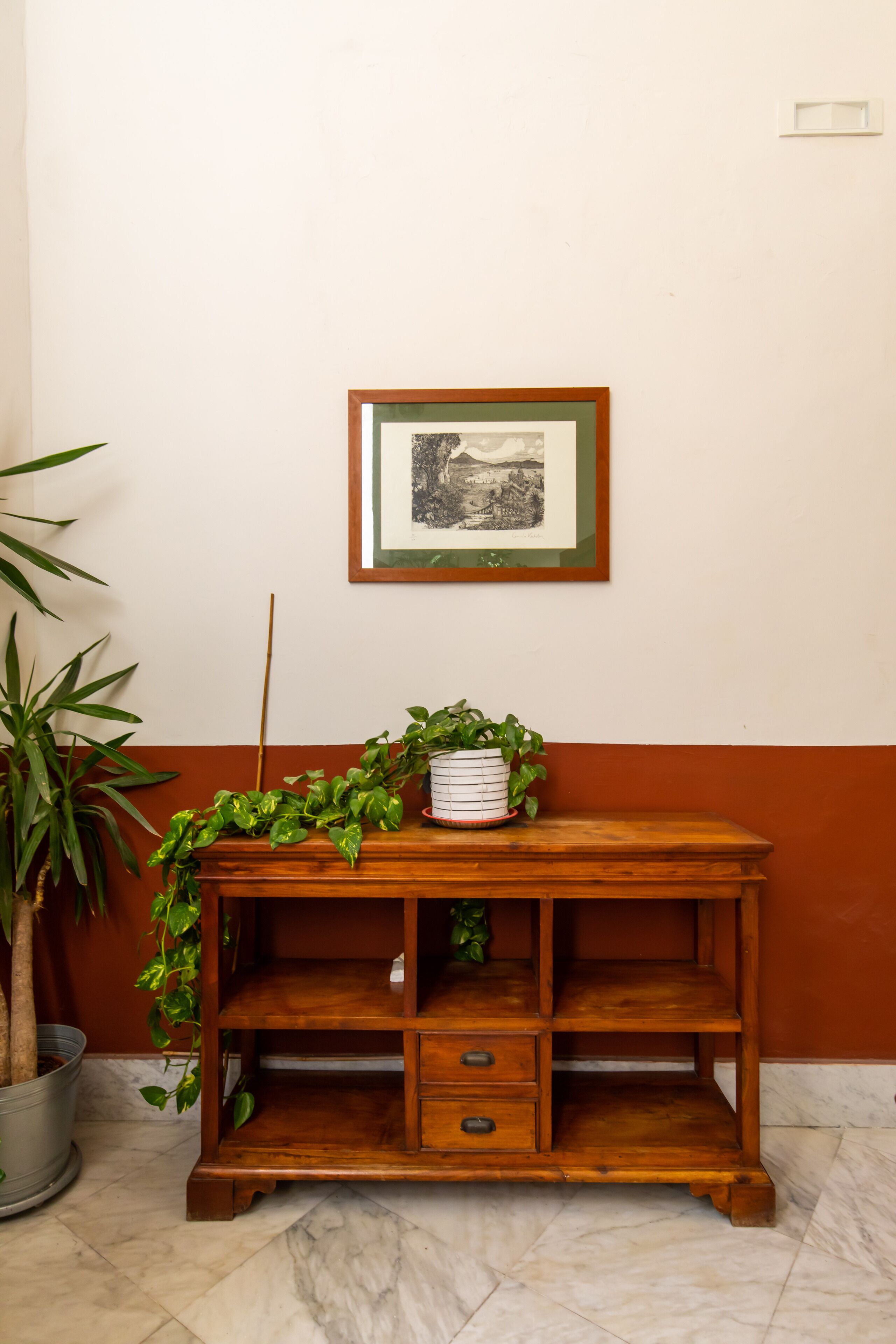 A wooden console table with a framed picture above it, a potted plant on the table, and a potted plant to the left.