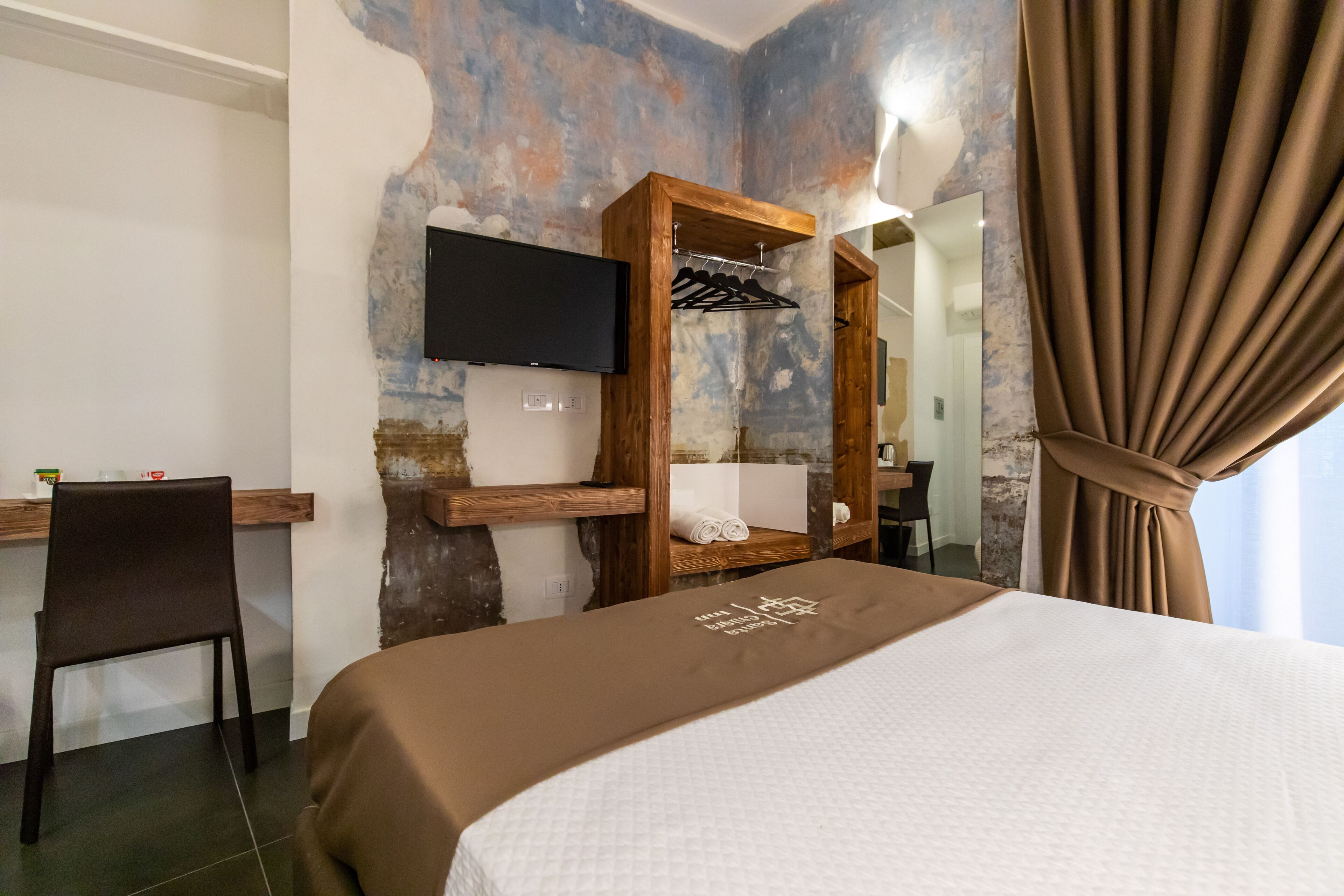 A hotel room with a bed, a TV mounted on the wall, a wooden shelf with towels, and a mirror.