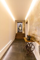 Un pasillo con una máquina de coser antigua sobre una mesa de madera, una escalera que lleva a una habitación con un espejo y un muro con textura.