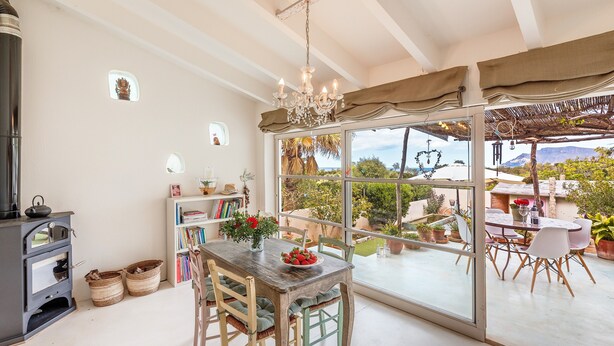 Une cuisine équipée d’une cuisinière, une table à manger avec des chaises, un lustre et une vue dégagée sur l’extérieur grâce à de grandes fenêtres.