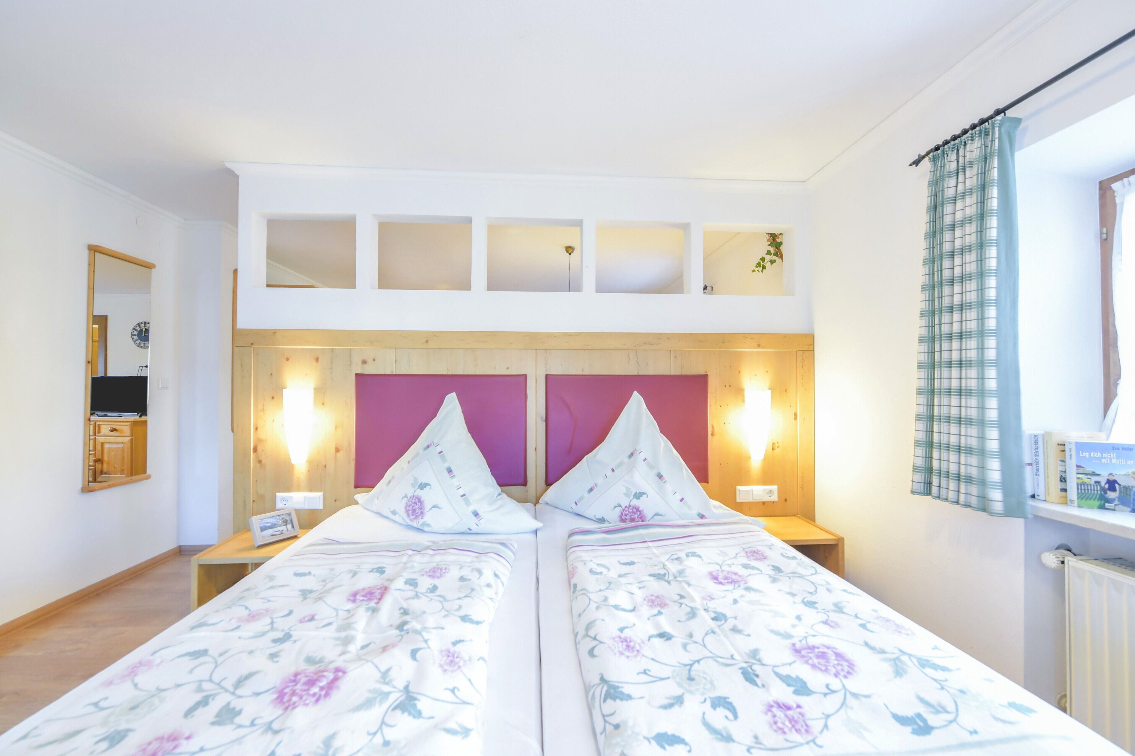 A double bed with floral bedding, a wooden headboard, two bedside lamps, a nightstand, a mirror, and a window with curtains.