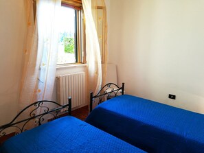 A room with two beds, a radiator, a window with curtains, and a wall socket.