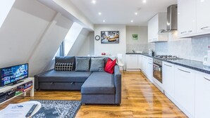 A modern apartment with a grey sofa, a flat-screen TV, a kitchen with white cabinets, and a dining area. - Luxurious Modern One Bedroom Apartment (Redhill)