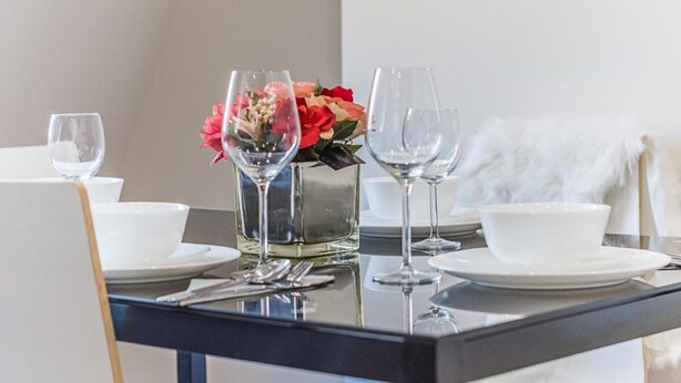A dining table set with white plates, clear wine glasses, and a vase with red and pink flowers. - Luxurious Modern One Bedroom Apartment (Redhill)
