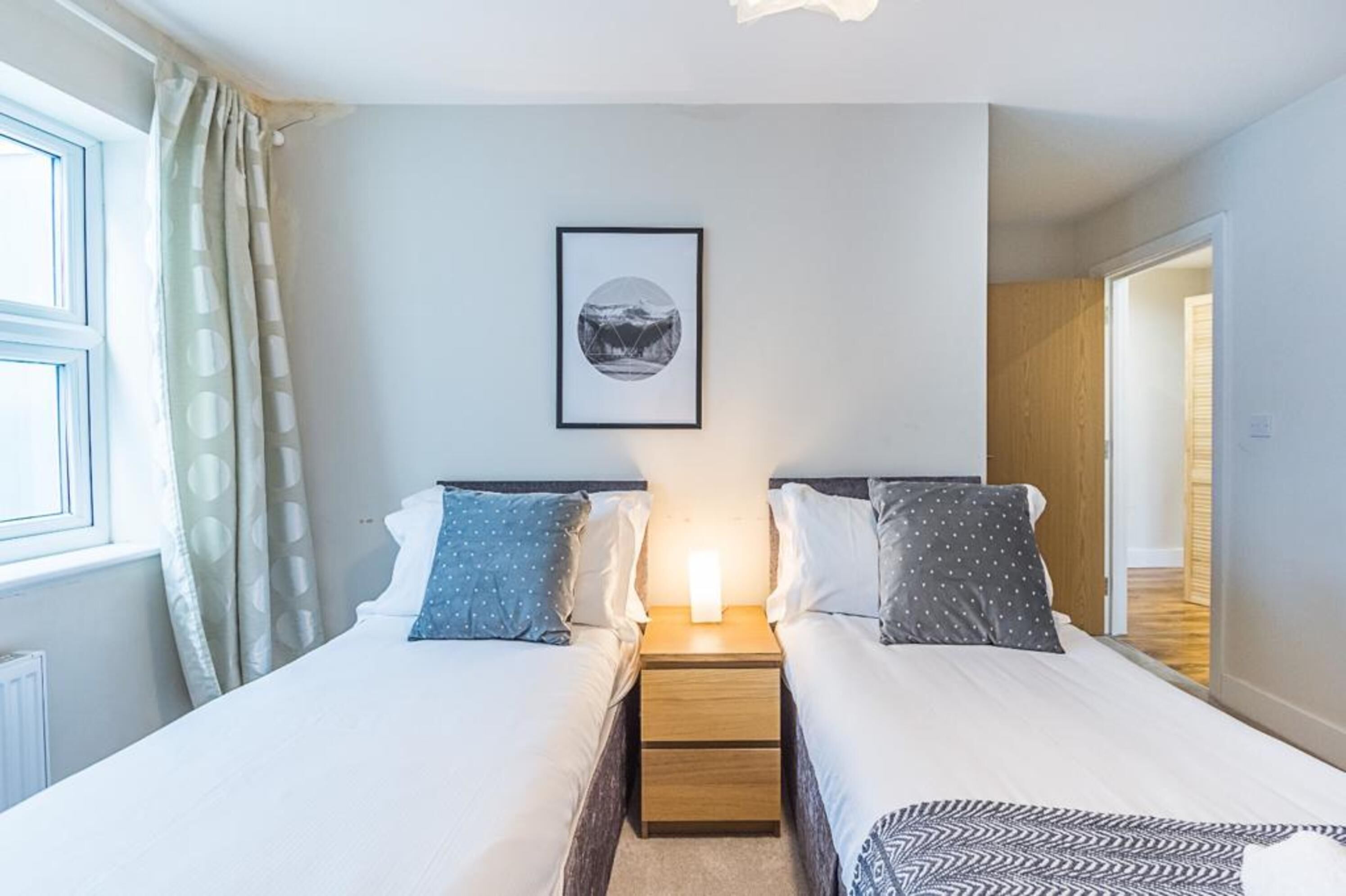 A bedroom with two beds, a bedside table with a lamp, a framed picture on the wall, and a window with curtains.