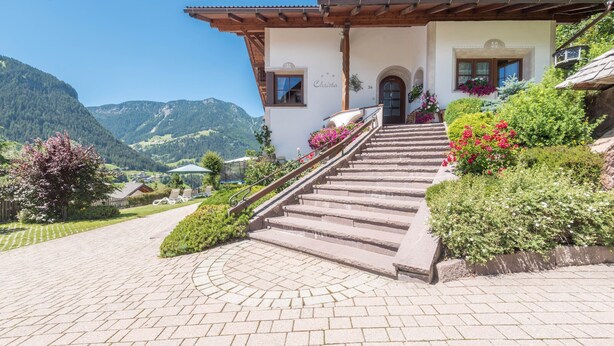 A building with a stone staircase leading to a door, surrounded by lush greenery and a mountainous backdrop. - Holiday Apartment "Christa B1" with Wi-Fi near Various Ski Resorts (Ortisei)