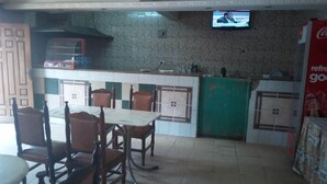 A restaurant with a tiled floor, wooden chairs, a table, a television mounted on the wall, and a Coca-Cola advertisement. - Mocharo Hotel (Isiolo)