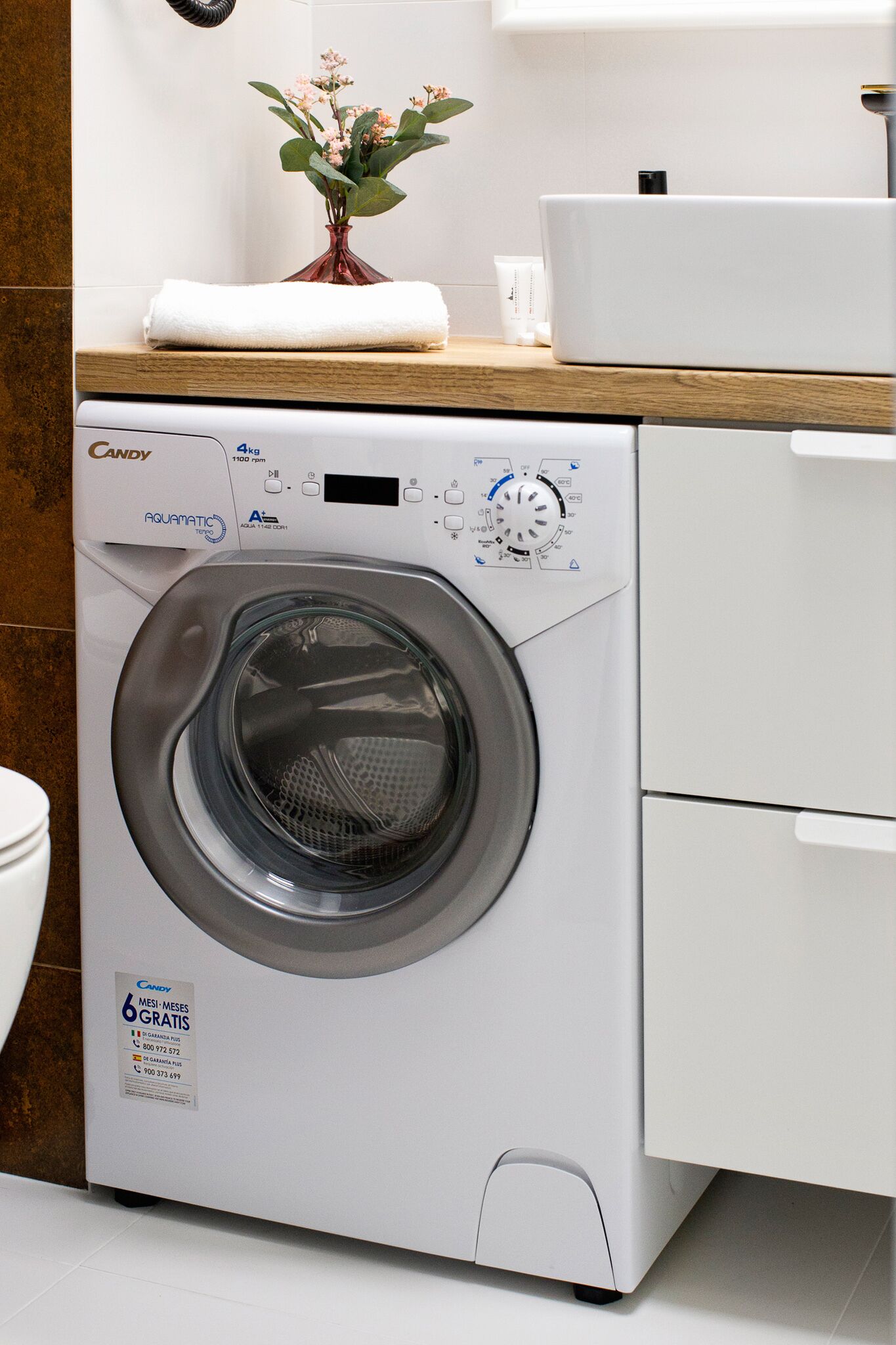 A front-loading washing machine with a control panel, a wooden countertop, and a vase with flowers.