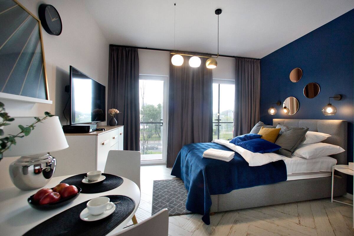 A modern hotel room with a large bed, a desk with a laptop, a TV, and a balcony with curtains.