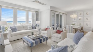 A spacious living room with a large window overlooking the ocean, a ceiling fan, and a dining area with a chandelier. - Stylish Beachfront Condo (Watersound)
