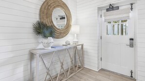 A white interior with a round mirror, a white console table with a plant, and a door with a window. - Stylish Beachfront Condo (Watersound)