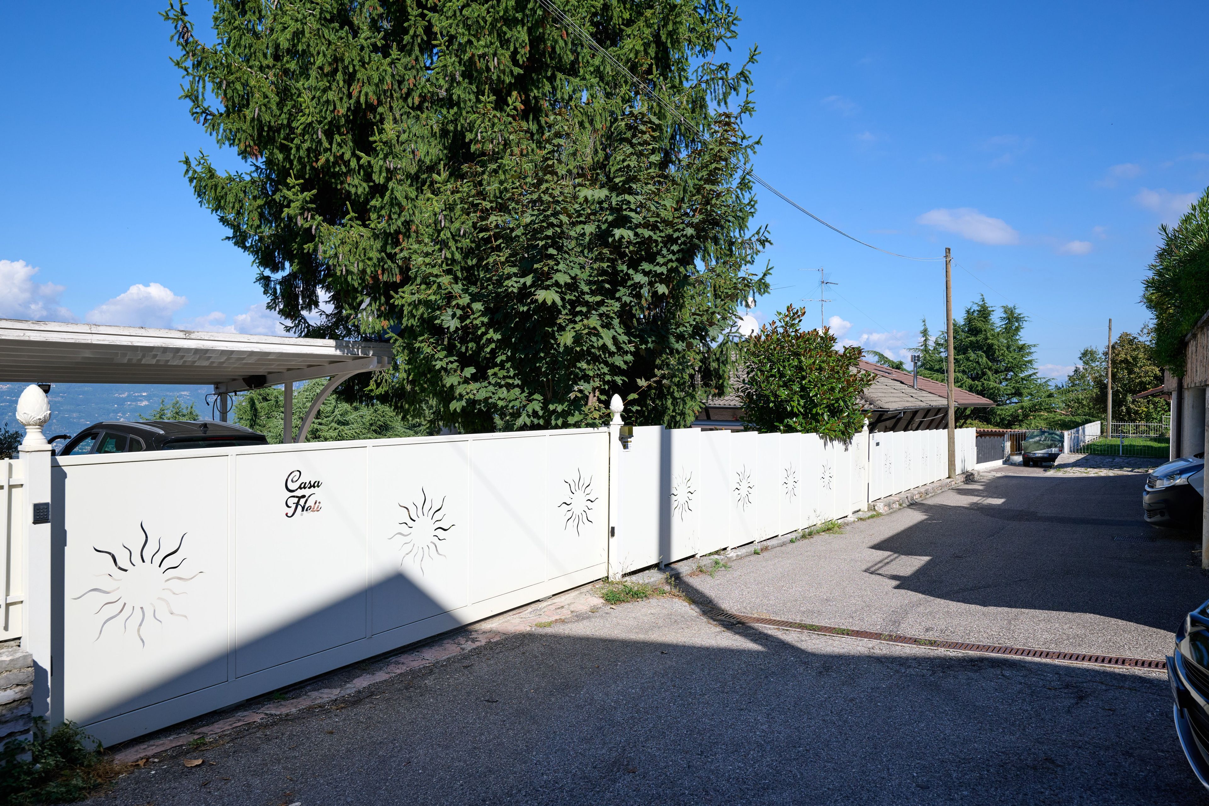 Una strada residenziale con un cancello bianco, un albero grande e auto parcheggiate.