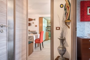 Une pièce avec une porte en bois, un coin repas avec une table et des chaises, un vase contenant des plantes séchées et un tableau encadré de rouge accroché au mur.