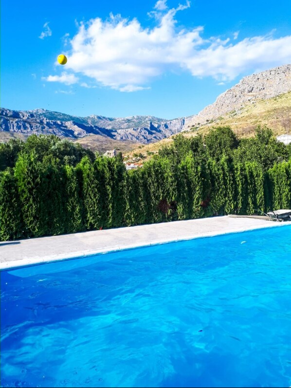 Une piscine à l’eau d’un bleu cristallin, entourée d’arbres verdoyants et dominée par un paysage montagneux en arrière-plan.