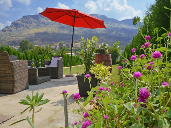 An outdoor seating area with wicker furniture and a red umbrella, surrounded by potted plants and flowers.