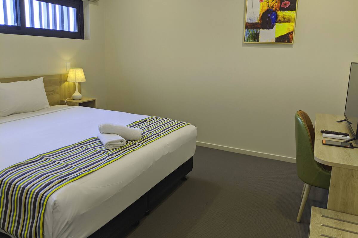 Habitación de hotel con cama, escritorio, silla, lámpara y un cuadro en la pared.