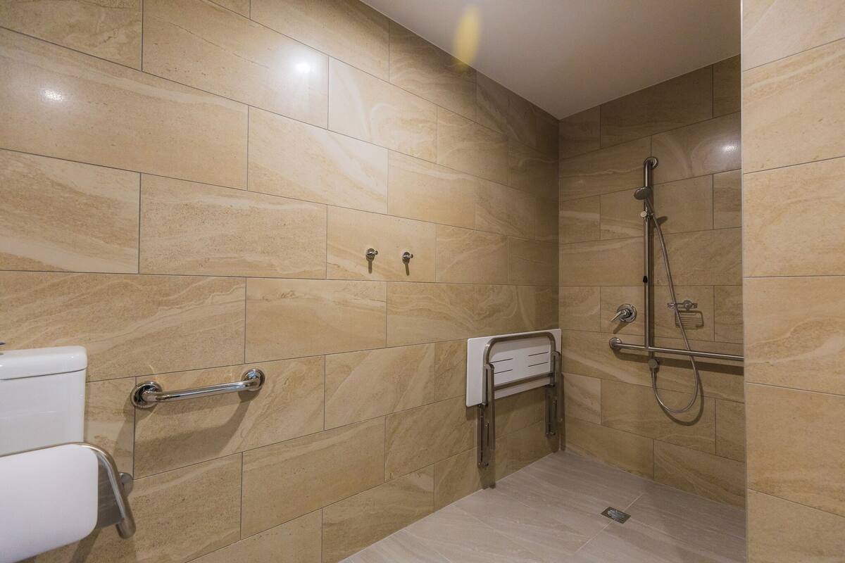 Un baño con baldosas de mármol color crema, un inodoro con barra de apoyo, un perchero y una ducha con cabezal de ducha manual.