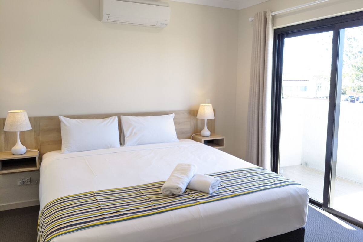 Habitación de hotel con una cama grande, dos lámparas de noche, un ventanal con cortinas y un equipo de aire acondicionado en la pared.