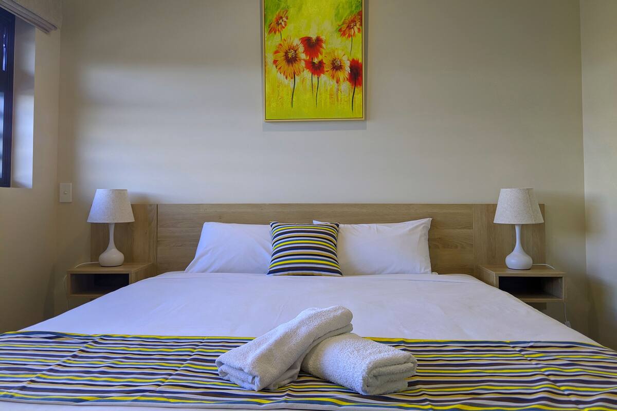 Una habitación de hotel con una cama grande, ropa de cama blanca, ropa de cama a rayas, un cuadro de flores en la pared y dos lámparas de noche.