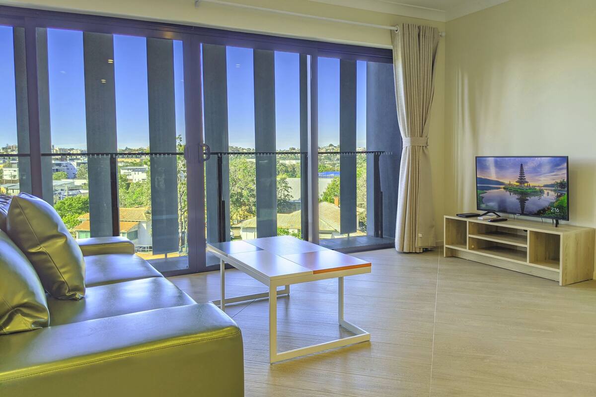 Una sala de estar moderna con un sofá, una mesa de centro, un televisor sobre un mueble de madera y un balcón con ventanas amplias.