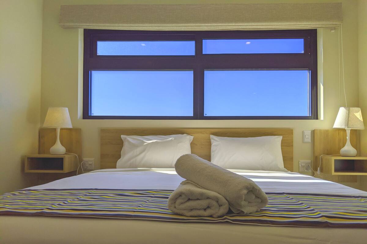 Una habitación de hotel con cama, dos mesitas de noche, una ventana y una toalla sobre la cama.
