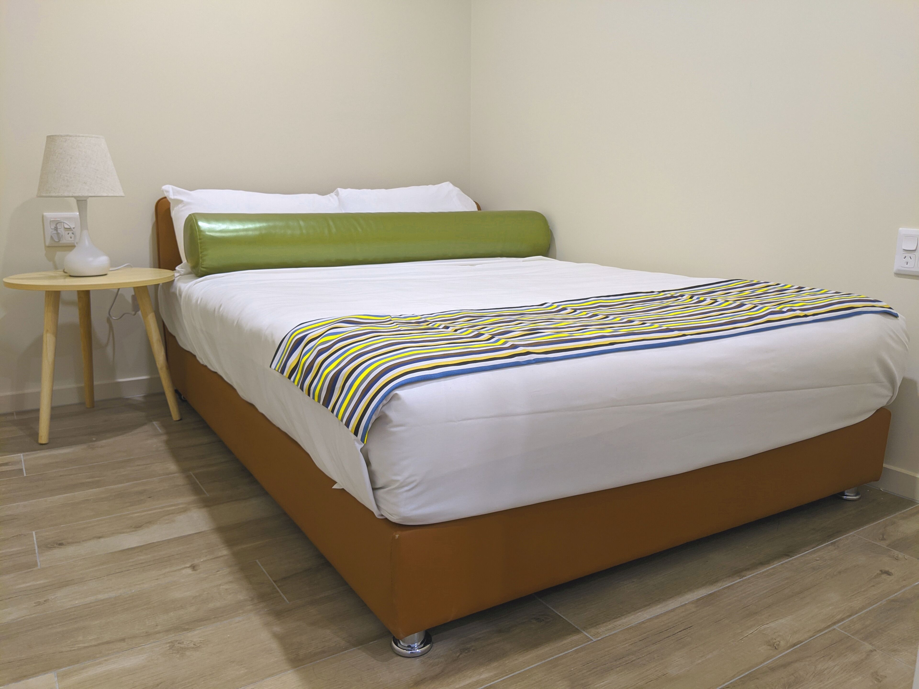 Un lit avec une tête de lit verte, un matelas blanc et une couverture à rayures. Une petite table de chevet en bois avec une lampe se trouve à côté du lit.