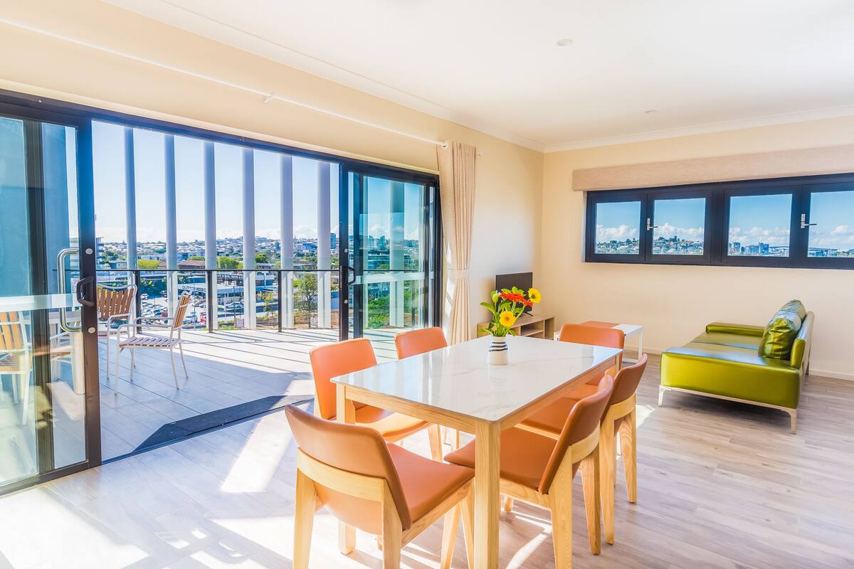 Una sala de estar moderna con balcón de vidrio, una zona de comedor con mesa y sillas, y un sofá verde.