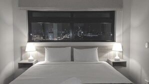 Habitación de hotel con una cama grande, dos mesitas de noche con lámparas y vista a la ciudad de noche.