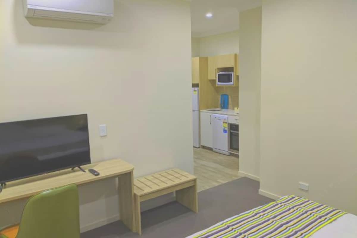 Habitación de hotel con cama, escritorio con televisor, pequeño banco y una kitchenette al fondo.