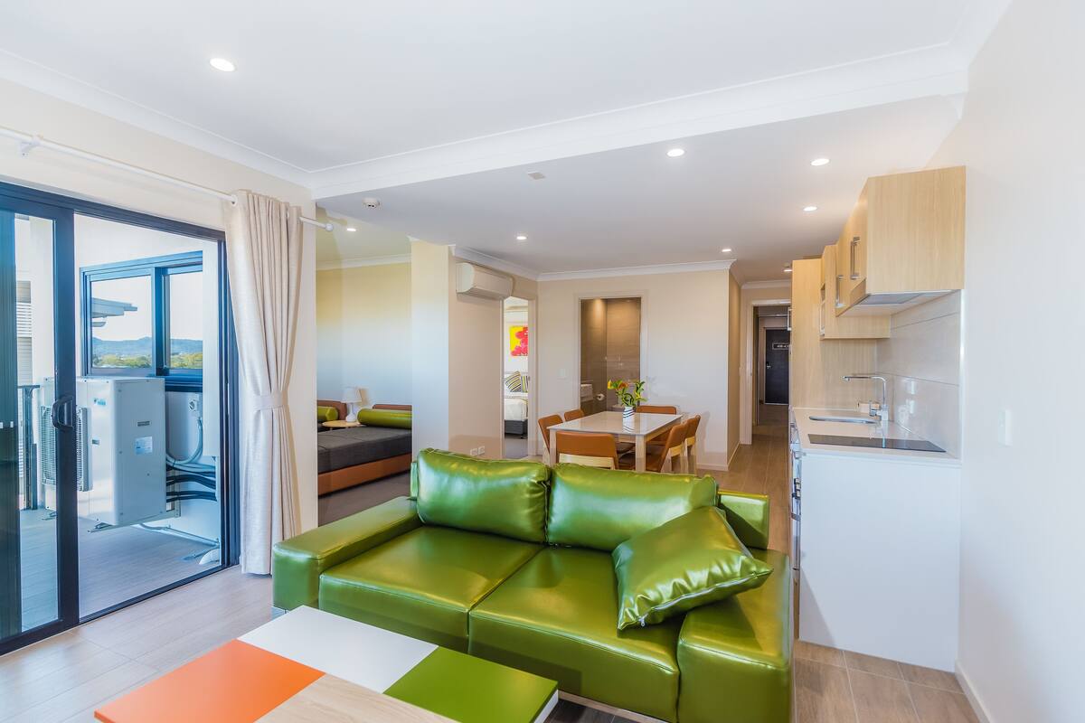 Una sala de estar moderna con un sofá de cuero verde, un comedor con una mesa y sillas de madera, y una cocina con gabinetes blancos.