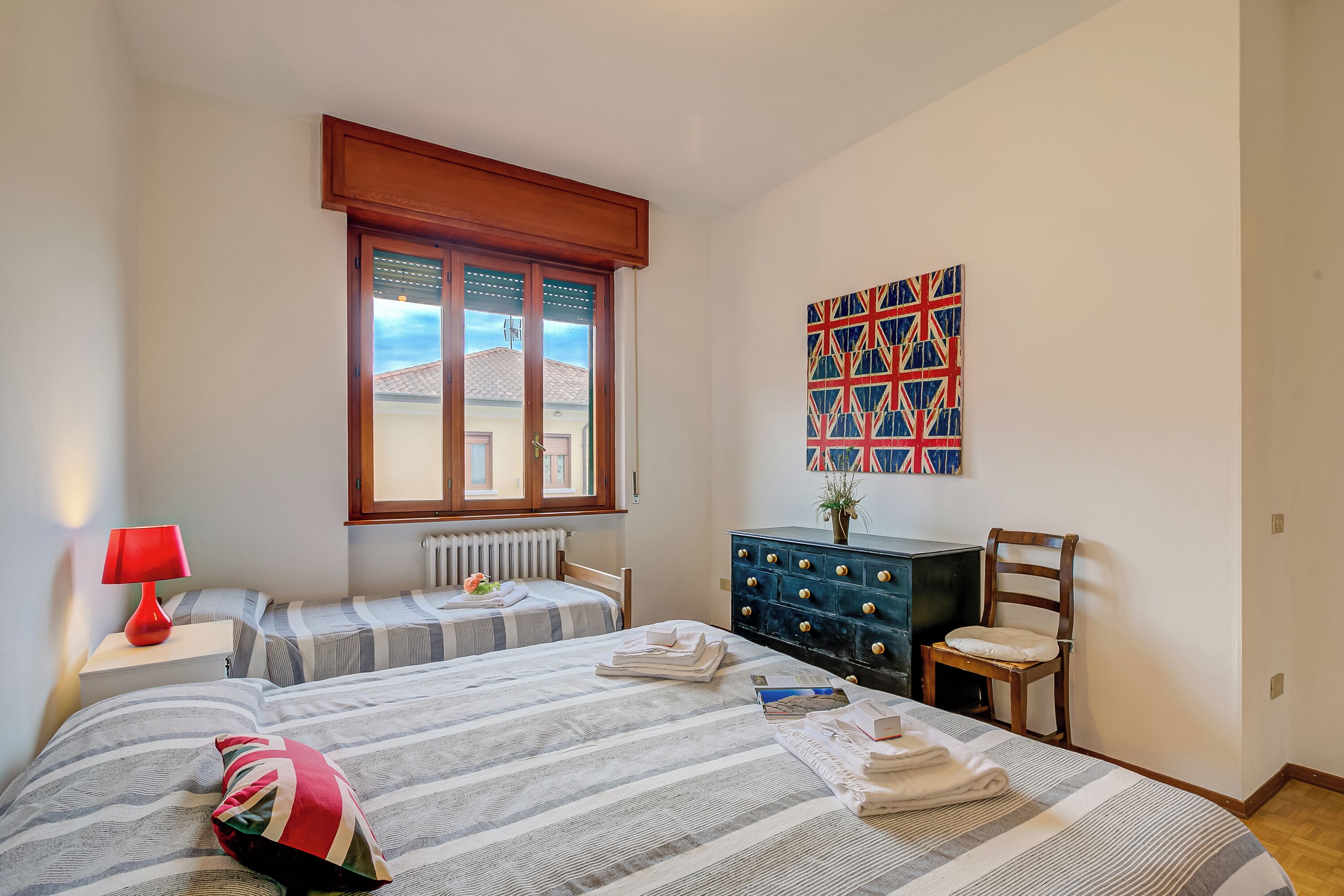 A bedroom with two beds, a dresser, a chair, a red lamp, and a window with blinds.